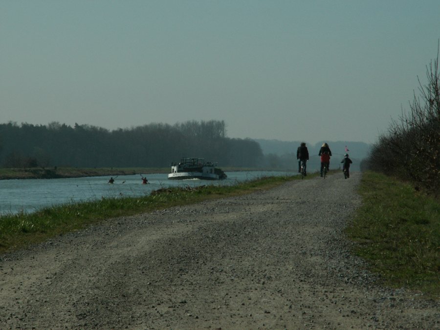 Drei Radfahrende auf einem Kiesweg an einem Fluss, daneben ein Ausflugsschiff und Kajaks.